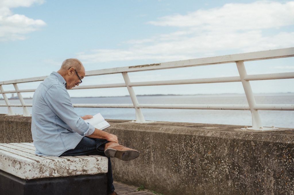 serious senior man reading book near fence on waterfront